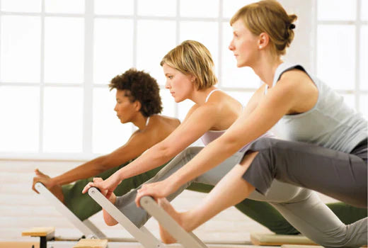 A group of people performing a synchronized Pilates reformer exercise, demonstrating proper form in a plank position while using the reformer machines in a bright, well-lit studio setting.